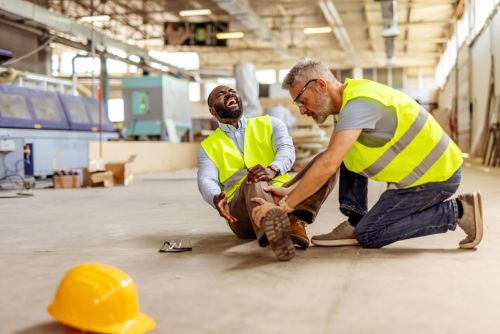 Two workers in an industrial workplace, wearing safety outfits, assisting after a workplace injury occurrence. Highlighting importance of safety protocols, hazard awareness, and emergency first aid response.
