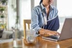 Unrecognizable woman with injured arm sitting at the table and using laptop at home.